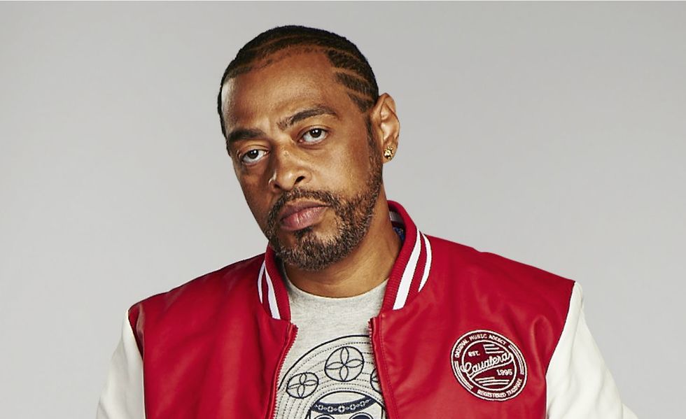 Thaíde, Homem com expressão séria, usando jaqueta vermelha e branca e camiseta cinza estampada, posa diante de fundo neutro, exibindo estilo urbano e confiante.