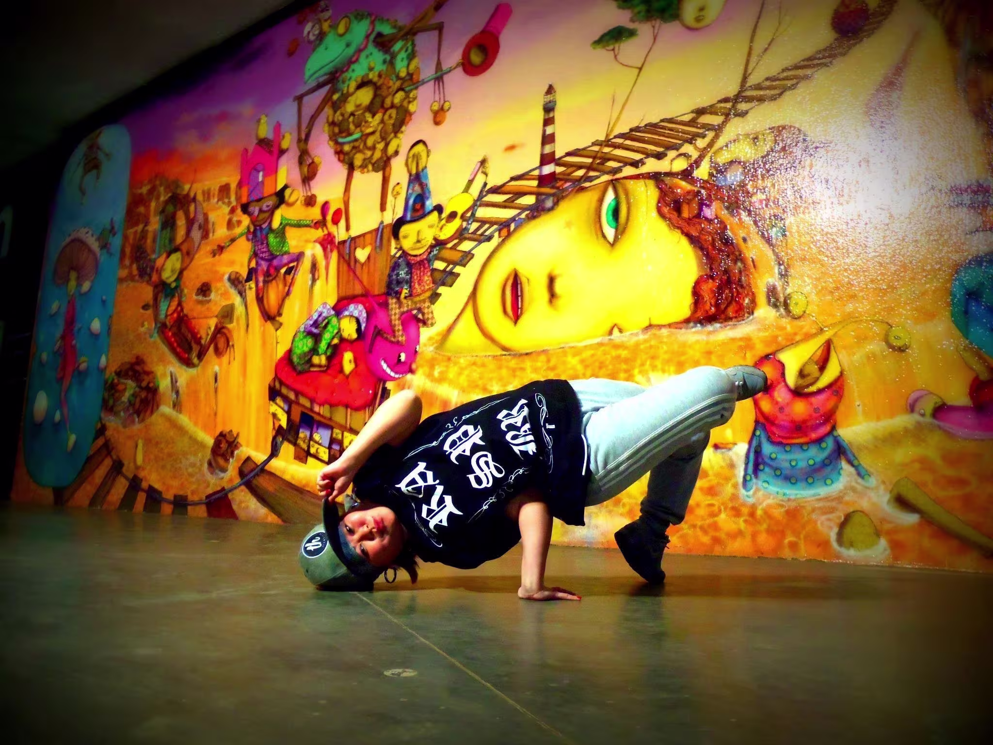 Miwa Kozuma, B-girl com boné cinza e camiseta preta faz uma pose de equilíbrio sobre uma mão em ambiente urbano, diante de um mural colorido com arte surrealista repleta de figuras e tons quentes.